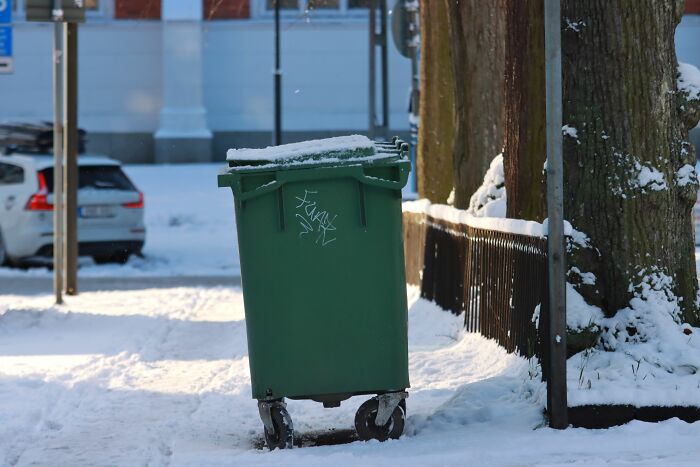 A green garbage can standing outdoors on a snowy day. Unethical Life Pro Tips.