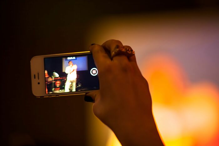 A hand holding a smartphone to record a live performance on screen, depicting unethical life pro tips