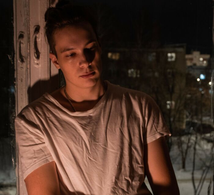 A young man sitting in dim lighting by a window, contemplating heavy secrets he keeps from everyone.