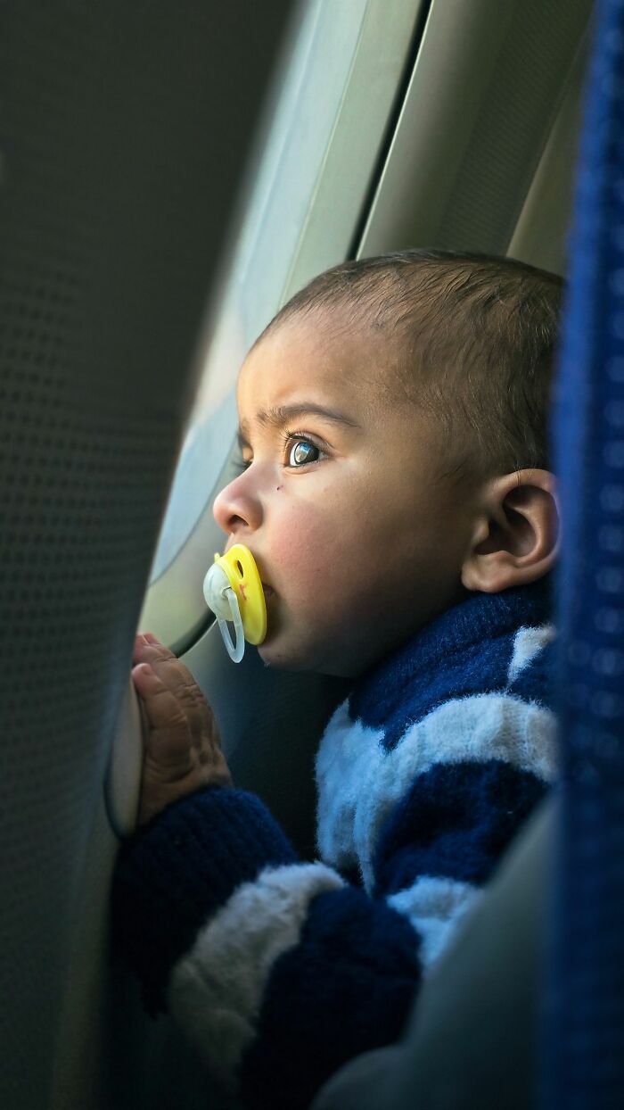 A baby with a pacifier looking out the window to get better seats on flights