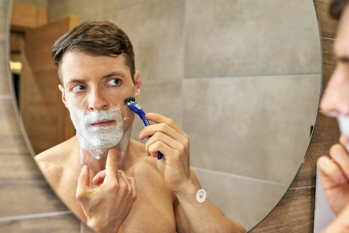 A young man shaving his face with foam in a bathroom mirror, demonstrating everyday clever life tricks for grooming.