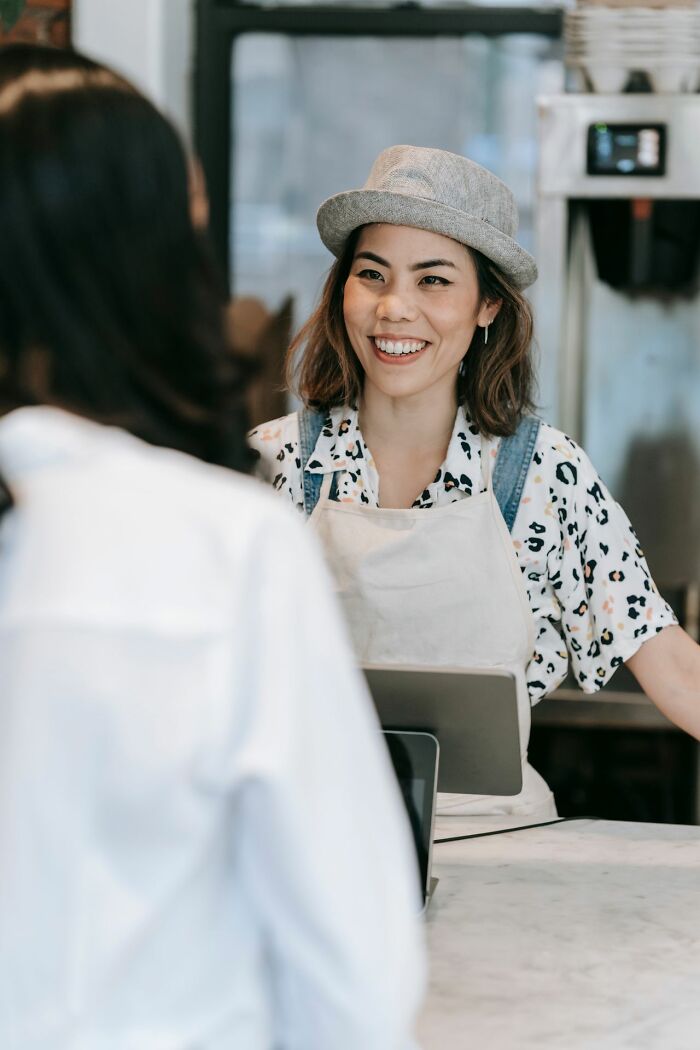 A smiling female barista wearing a hat and apron at a coffee shop counter, representing clever life tricks in service.