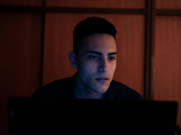 A young man stares intently at his glowing laptop screen in a dark room, searching online forums.