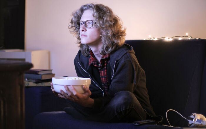 A person with glasses holding a bowl on a couch, pondering personal life secrets while watching TV.