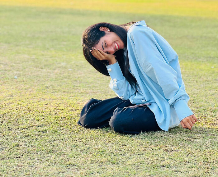 A young woman laughing embarrassingly while sitting on the grass outdoor.