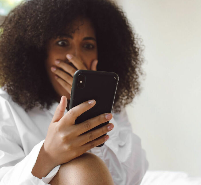 Surprised woman covering her mouth while looking at her smartphone during funny instances.