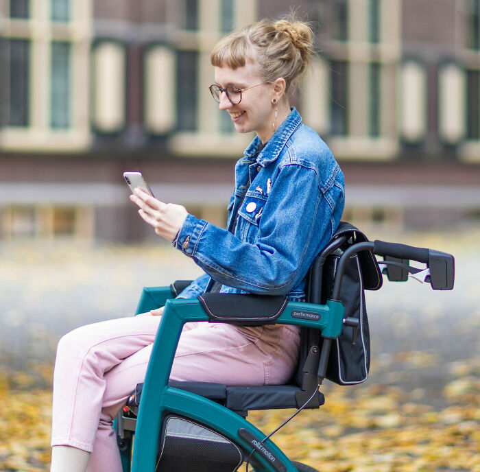 A young woman in glasses and denim jacket smiling while using a wheelchair mobility aid outdoors
