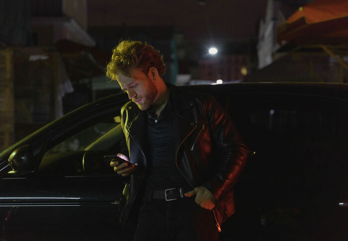 A man in a leather jacket stands by a car at night, representing a manosphere adherent looking at his phone.