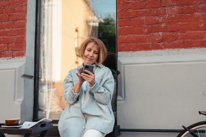 Middle aged woman sitting outdoors and looking at her smartphone during funny instances.
