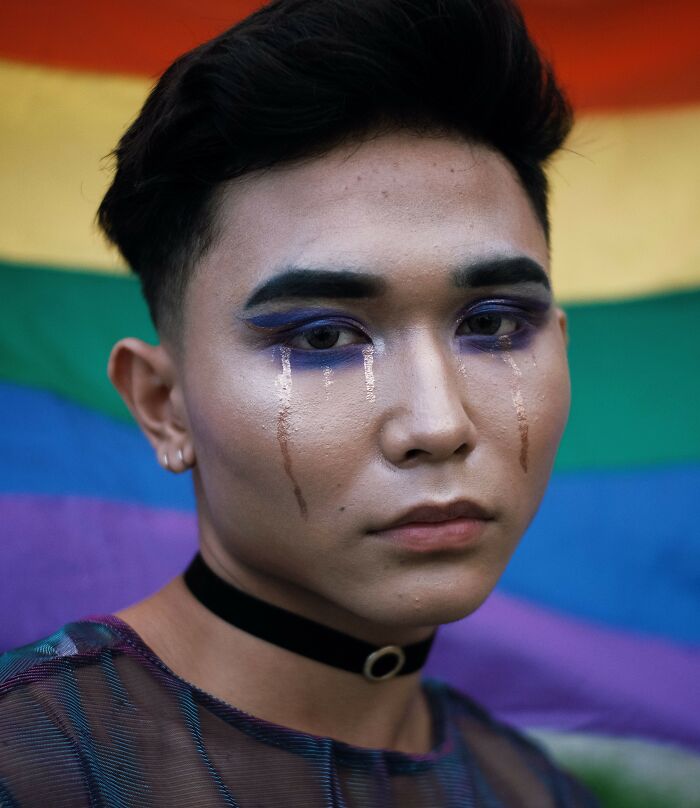 A person with makeup and painted tears standing in front of a pride flag carrying personal secrets.