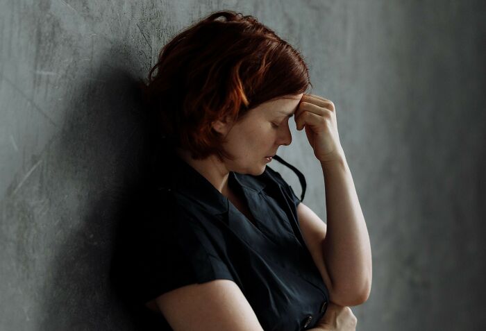A red-haired woman leaning against a wall with her hand on her forehead, stressed about secret burdens.