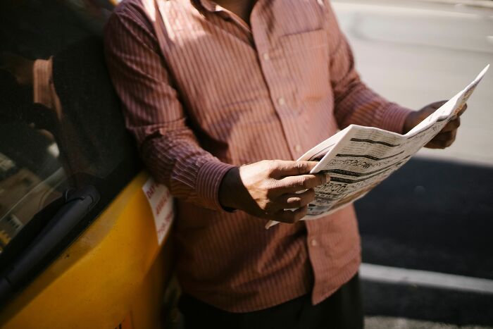 Man reading a newspaper beside a yellow car, catching up on tiny communities drama