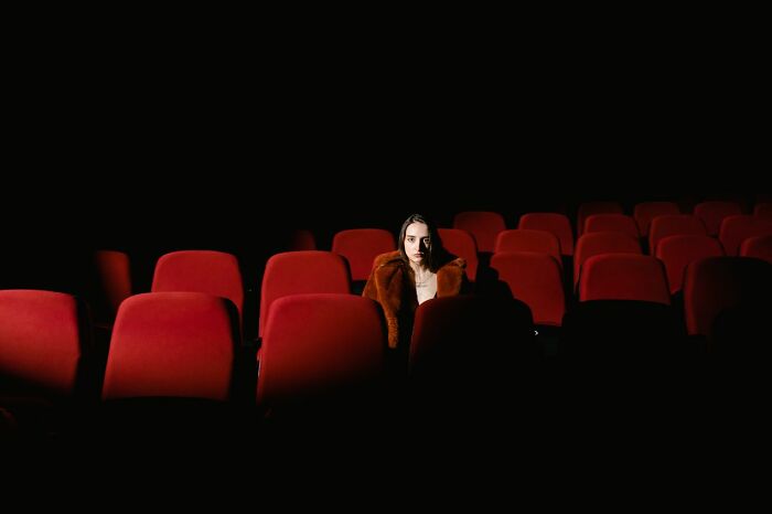 Woman sitting alone in an empty dark movie theater showing red seats, illustrating funny instances.