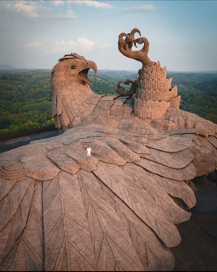 A man standing on the wing of a colossal bird statue, an entertaining pic from a scenic mountain overview