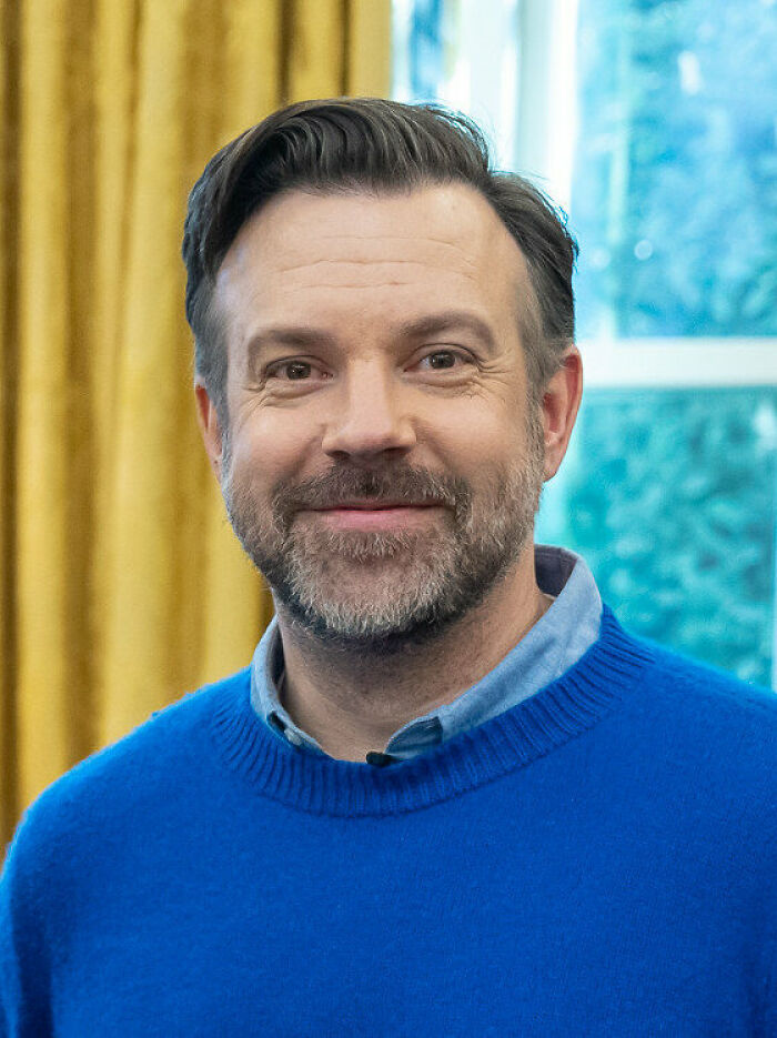 Jason Sudeikis smiling in a blue sweater, an embodiment of positive masculinity