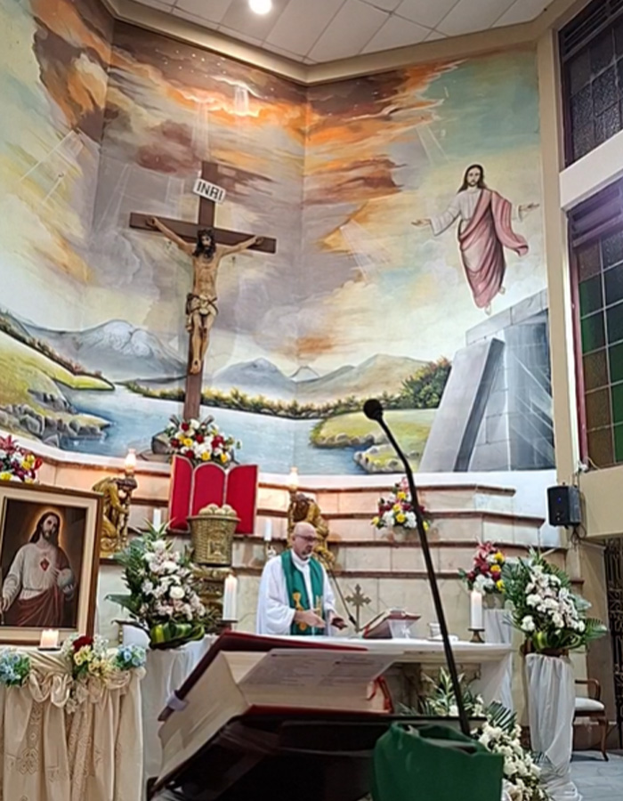 Priest standing at altar with large crucifix and flower arrangements