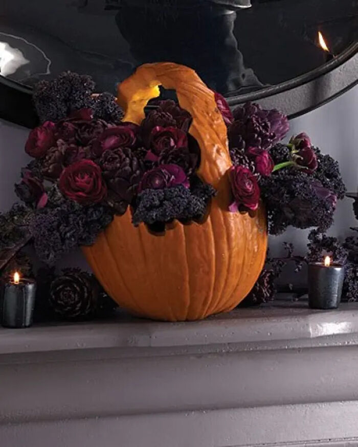 A carved pumpkin flower basket filled with dark purple flowers and kale for spooky Halloween homes.