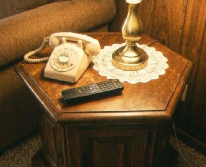 Nostalgic 80s photo showing a vintage beige rotary telephone and TV remote on a wooden end table.