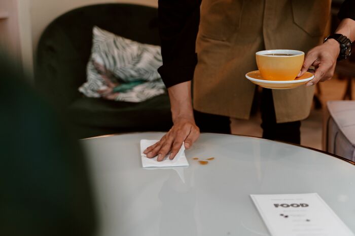 A waiter holding a cup of coffee while wiping a spilled stain on a table.