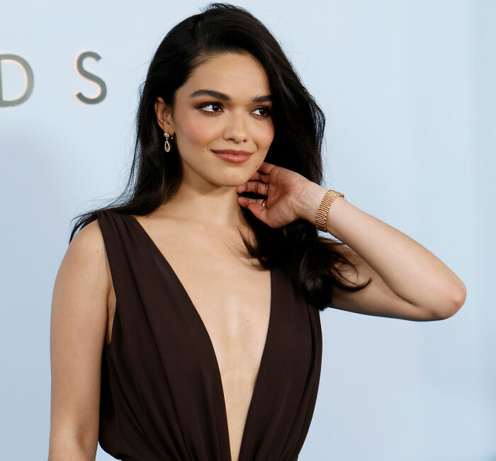 Actress Rachel Zegler posing in a brown V-neck dress on a light blue background.