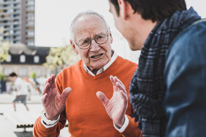 An older man wearing an orange sweater talks animatedly, gesturing with his hands during a discussion. An older man wearing an orange sweater talks animatedly, gesturing with his hands during a discussion.