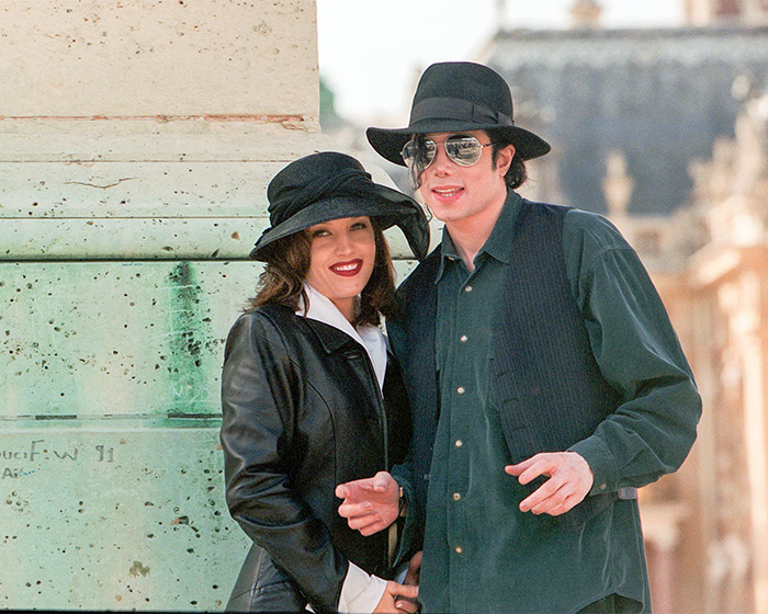 Michael Jackson and Lisa Marie Presley posing together outdoors wearing black hats and dark coats.