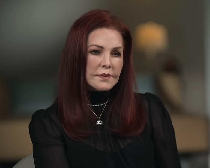 Priscilla Presley sitting indoors wearing a black top and Chanel necklace.