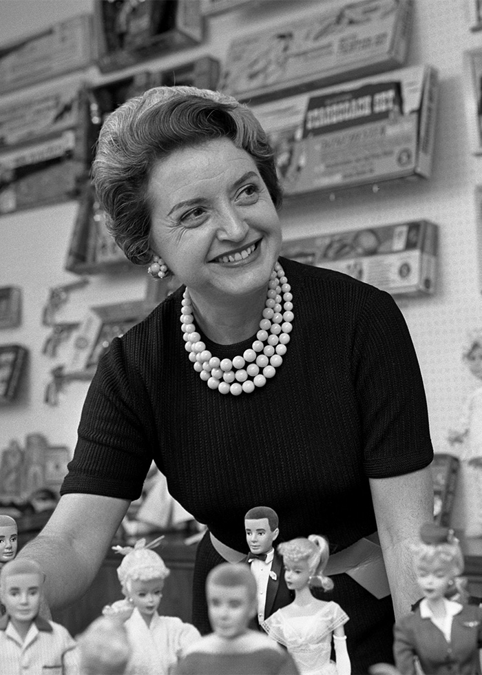 Black and white portrait of Mattel co-founder Ruth Handler standing behind vintage Barbie dolls.