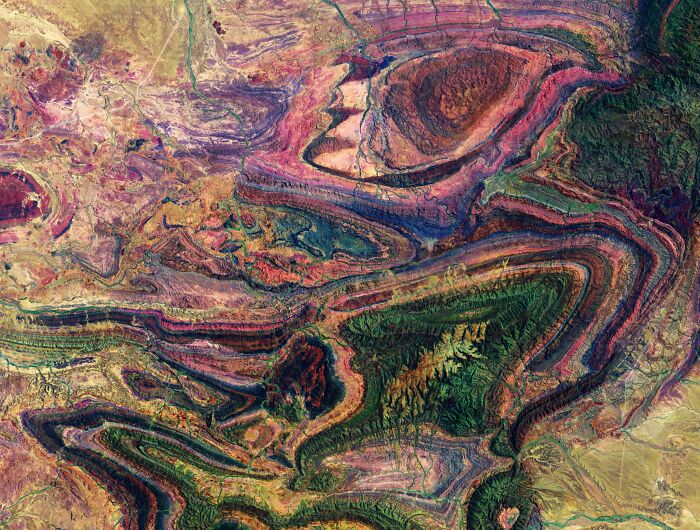 A surreal satellite photo displaying vibrant, colorful geological rock formations and curved strata from above.