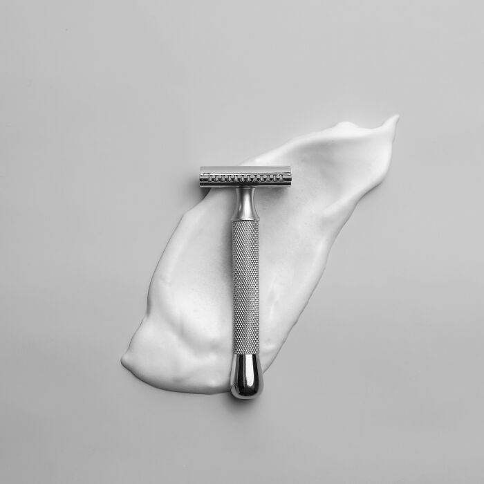 A classic metal safety razor placed over white shaving cream on a gray background