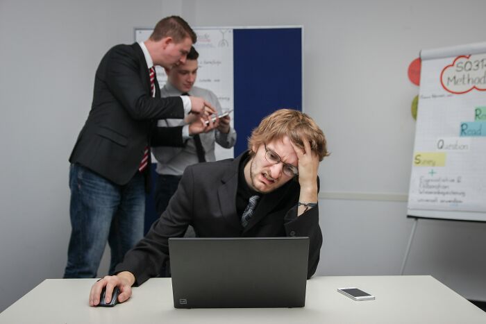 A stressed man working at a laptop while colleagues talk, illustrating how professionals lose trust.