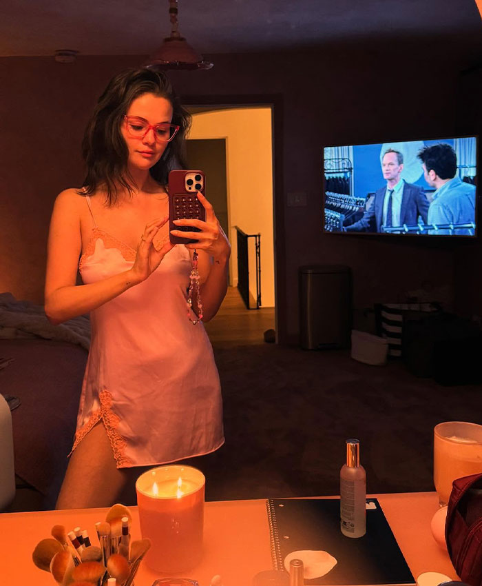 Selena Gomez taking a mirror selfie wearing glasses and a pink nightgown in a dimly lit room.