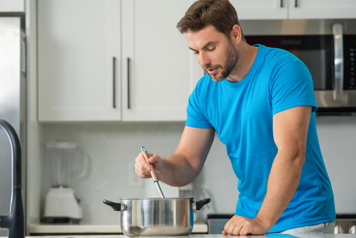 A young man in a blue t-shirt cooks and stirs a pot in a bright kitchen, showcasing life living with guys.