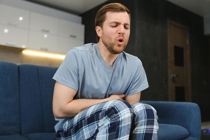 A man in plaid pants sitting on a blue sofa holds his stomach, a surprising aspect of living with guys.