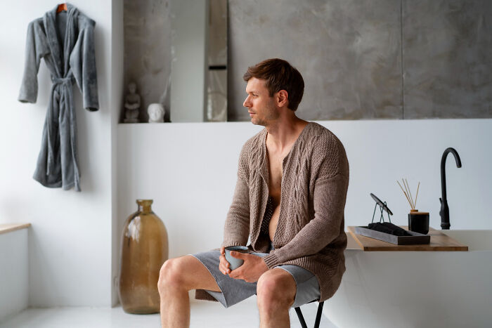 A thoughtful man sitting in a bathroom with a mug, reflecting on the realities of living with guys.