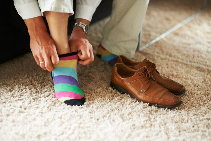 A man putting on colorful striped socks next to his dress shoes, showing quirks of living with guys.