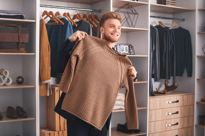 A smiling man holds up a brown sweater in a walk-in closet, a detail of living with guys.