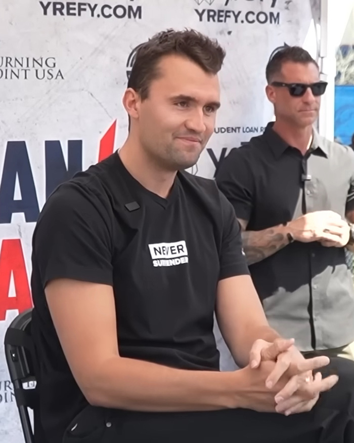 Charlie Kirk seated in a black shirt while attending a public event.