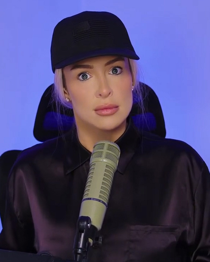 Influencer posing with wide eyes in a cap at a microphone, wearing a parody Erika Kirk Halloween costume.