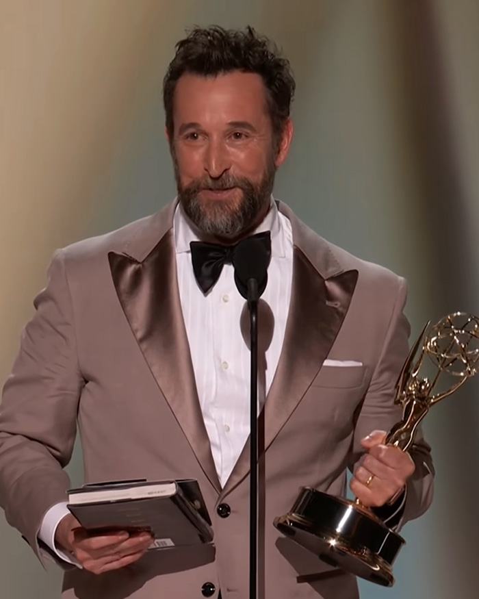 Noah Wyle from The Pitt holding an award on stage in a suit. Noah Wyle from The Pitt holding an award on stage in a suit.