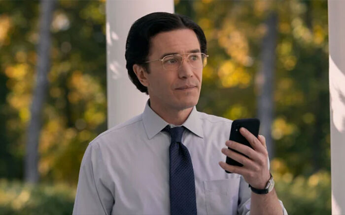 Man wearing a shirt, tie, and wire-rimmed glasses holding a mobile phone in a Netflix miniseries scene