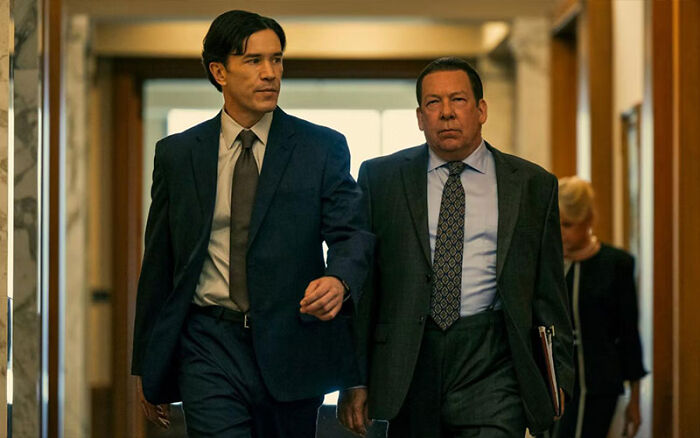 Two men in formal suits walking down a hallway in a Netflix miniseries scene.