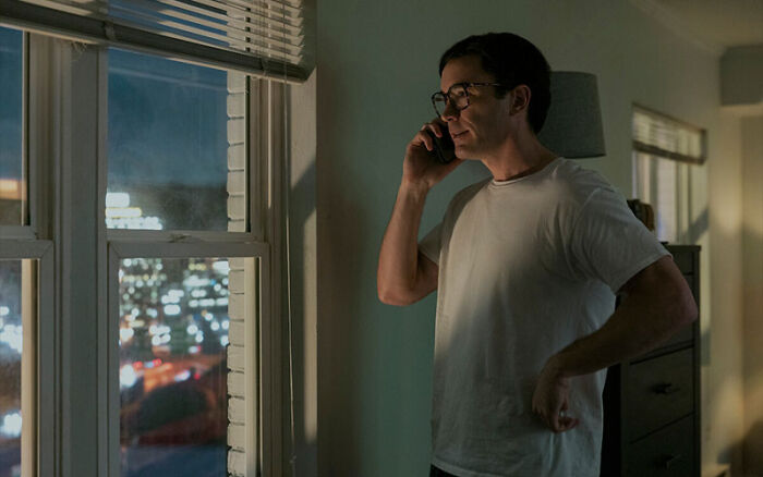 Man wearing glasses and a white t-shirt talking on a smartphone near a window in a Netflix miniseries