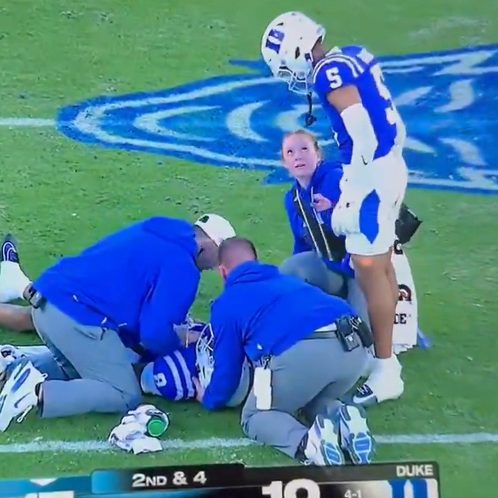 Football player and woman in blue standing by teammate receiving treatment
