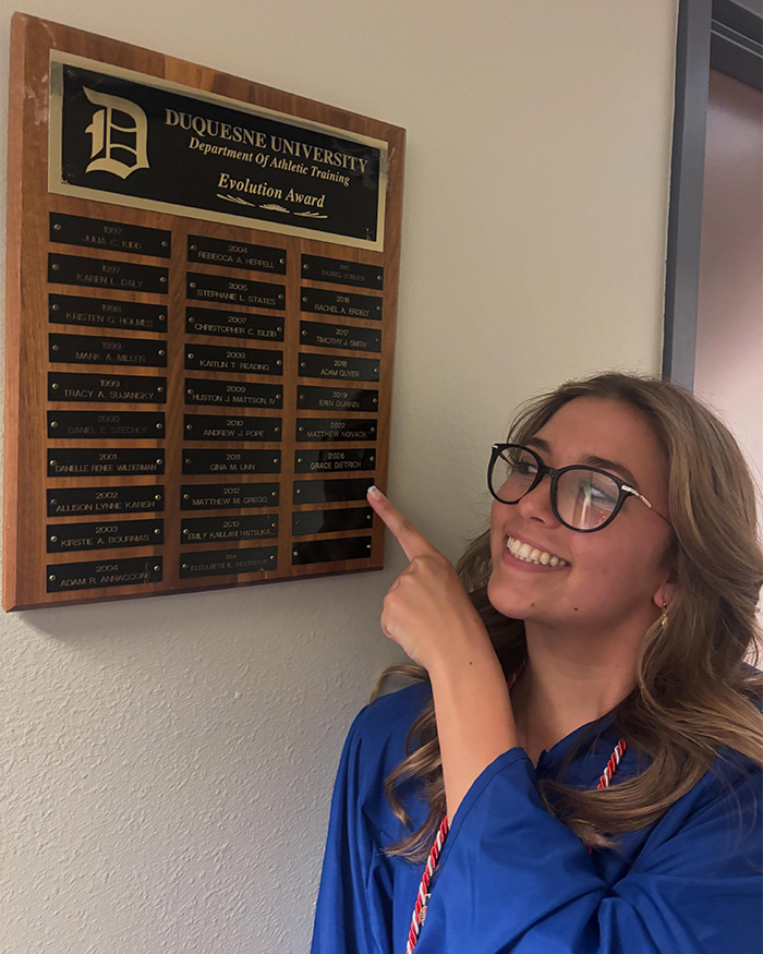 Woman in graduation gown pointing to Duquesne University athletic award plaque