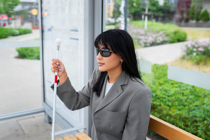 A visually impaired woman wearing sunglasses with a white cane waiting at a bus stop with quick-fire jokes