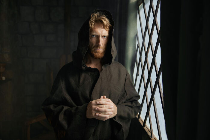 Red-bearded man in a dark hood standing near a window, illustrating quick-fire jokes.