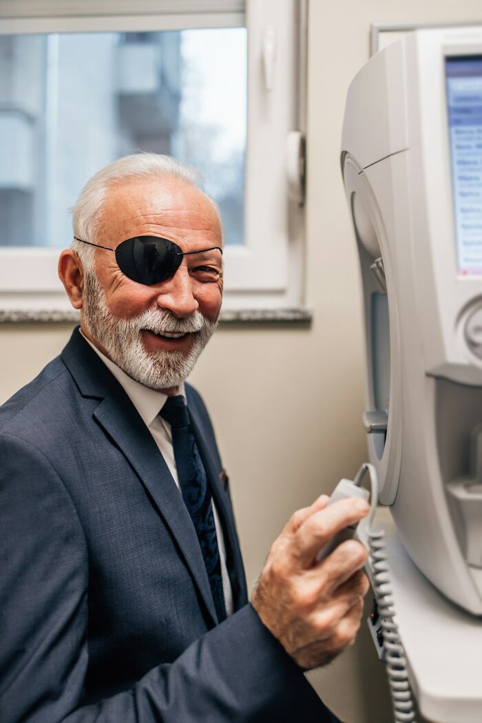 An older man with an eye patch smiling near an eye exam machine, sharing true stories from his life.