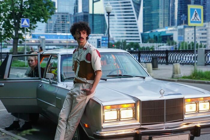 A man with a mustache leaning on a silver vintage car in the city, part of true stories people tell.