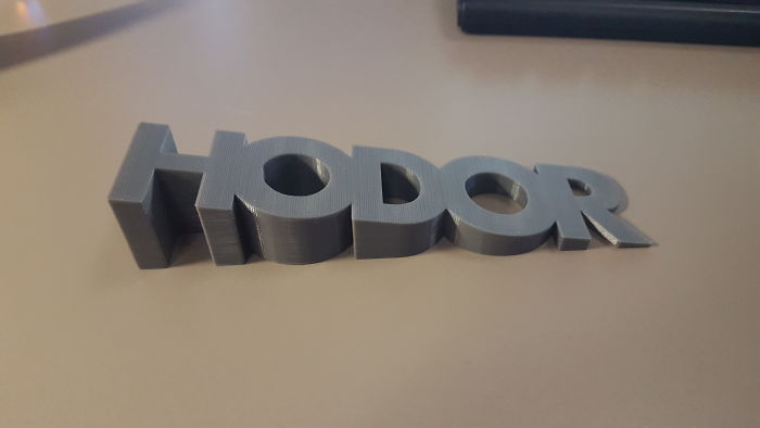 A grey 3D printer plastic door stopper spelling the word Hodor sitting on a desk